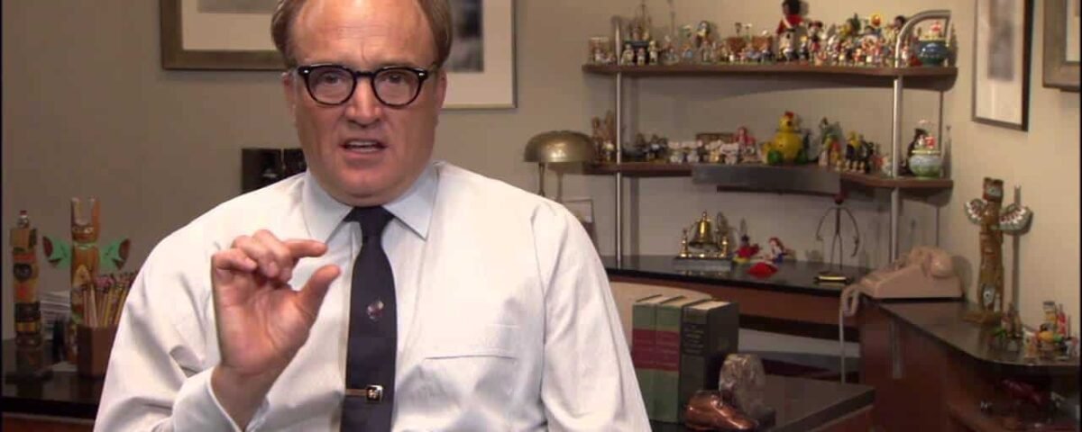 Un homme à lunettes portant une chemise blanche et une cravate fait des gestes avec sa main alors qu'il est assis dans un bureau rempli de figurines colorées et de livres liés à Don DaGradi en arrière-plan.