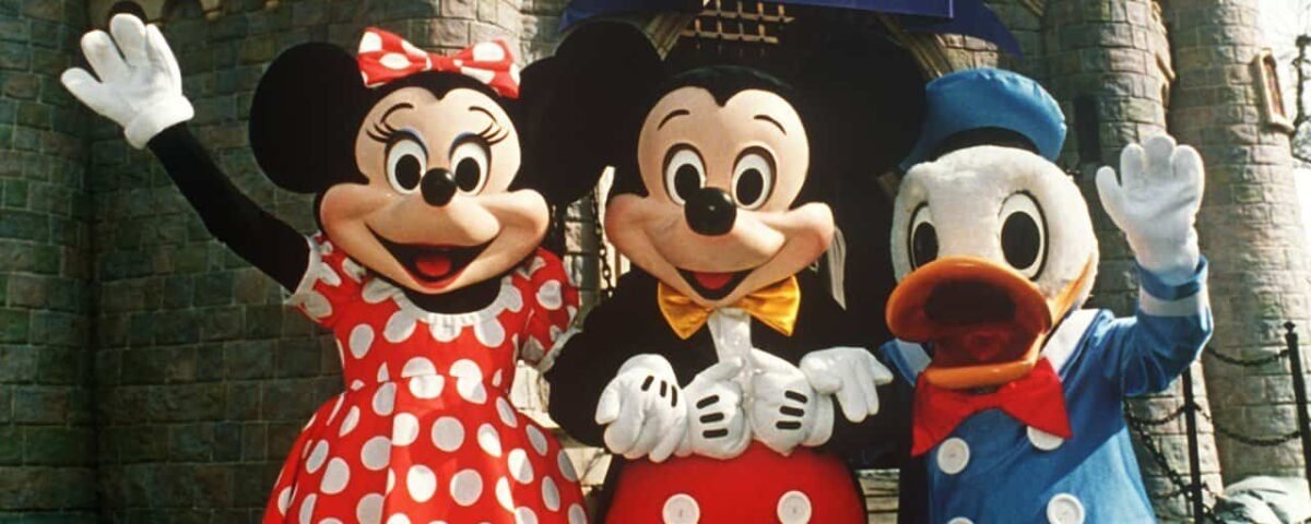 Minnie Mouse, Mickey Mouse et Donald Duck posant joyeusement devant un château d'Euro Disneyland. Minnie porte une robe rouge à pois, Mickey en tenue classique et Donald en