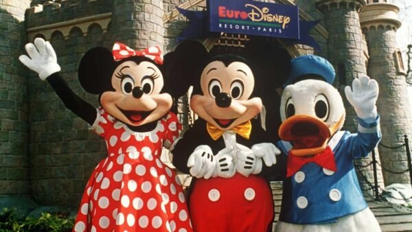 Minnie Mouse, Mickey Mouse et Donald Duck posant joyeusement devant un château d'Euro Disneyland. Minnie porte une robe rouge à pois, Mickey en tenue classique et Donald en