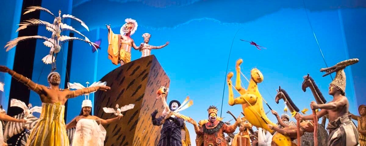 Une représentation théâtrale vibrante du « Roi Lion » mettant en vedette des acteurs vêtus de costumes élaborés représentant des animaux et des créatures mythiques sur une scène avec un fond bleu vif et des poses dynamiques.