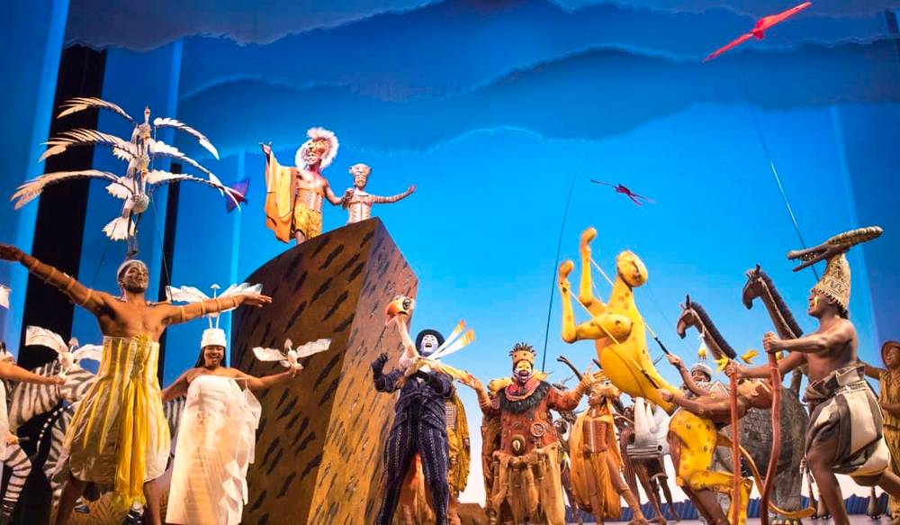 Une représentation théâtrale vibrante du « Roi Lion » mettant en vedette des acteurs vêtus de costumes élaborés représentant des animaux et des créatures mythiques sur une scène avec un fond bleu vif et des poses dynamiques.