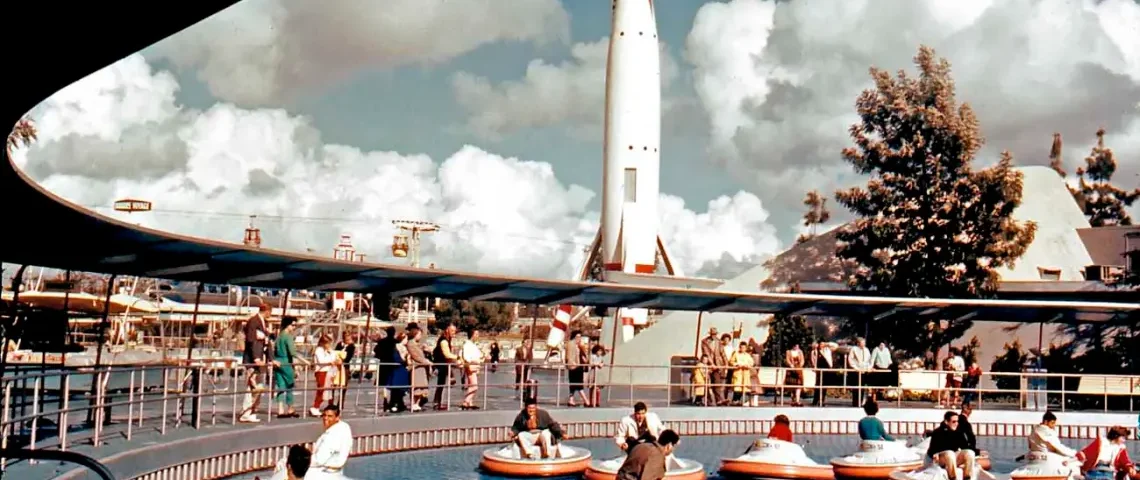 Photo couleur vintage de personnes dans des bateaux tamponneurs dans une piscine circulaire, avec une grande soucoupe volante les surplombant, sur fond de ciel bleu et de nuages duveteux.