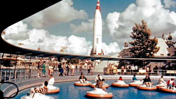 Photo couleur vintage de personnes dans des bateaux tamponneurs dans une piscine circulaire, avec une grande soucoupe volante les surplombant, sur fond de ciel bleu et de nuages duveteux.