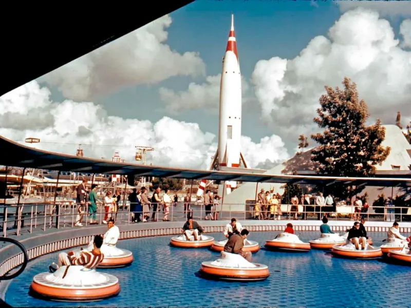Photo couleur vintage de personnes dans des bateaux tamponneurs dans une piscine circulaire, avec une grande soucoupe volante les surplombant, sur fond de ciel bleu et de nuages duveteux.