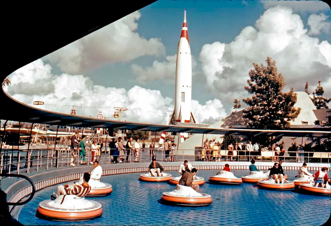 Photo couleur vintage de personnes dans des bateaux tamponneurs dans une piscine circulaire, avec une grande soucoupe volante les surplombant, sur fond de ciel bleu et de nuages duveteux.