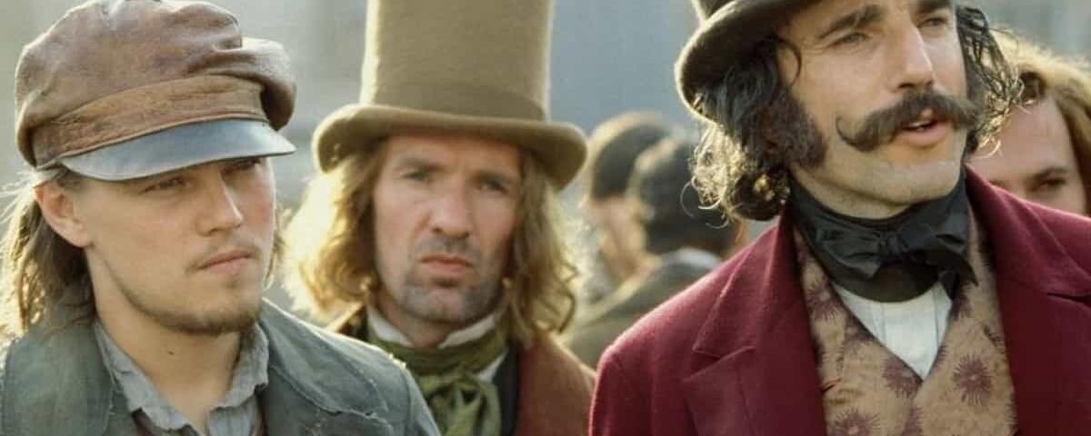 Trois hommes en tenue historique, deux portant des chapeaux hauts et un avec une casquette, se tiennent ensemble, représentant les personnages du film d'époque "Gangs of New York". L'homme au centre arbore un