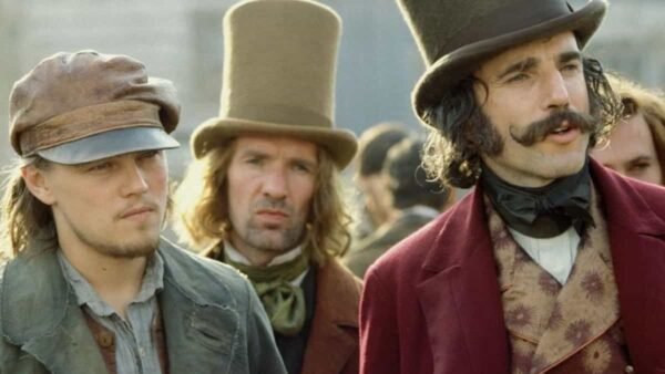Trois hommes en tenue historique, deux portant des chapeaux hauts et un avec une casquette, se tiennent ensemble, représentant les personnages du film d'époque "Gangs of New York". L'homme au centre arbore un