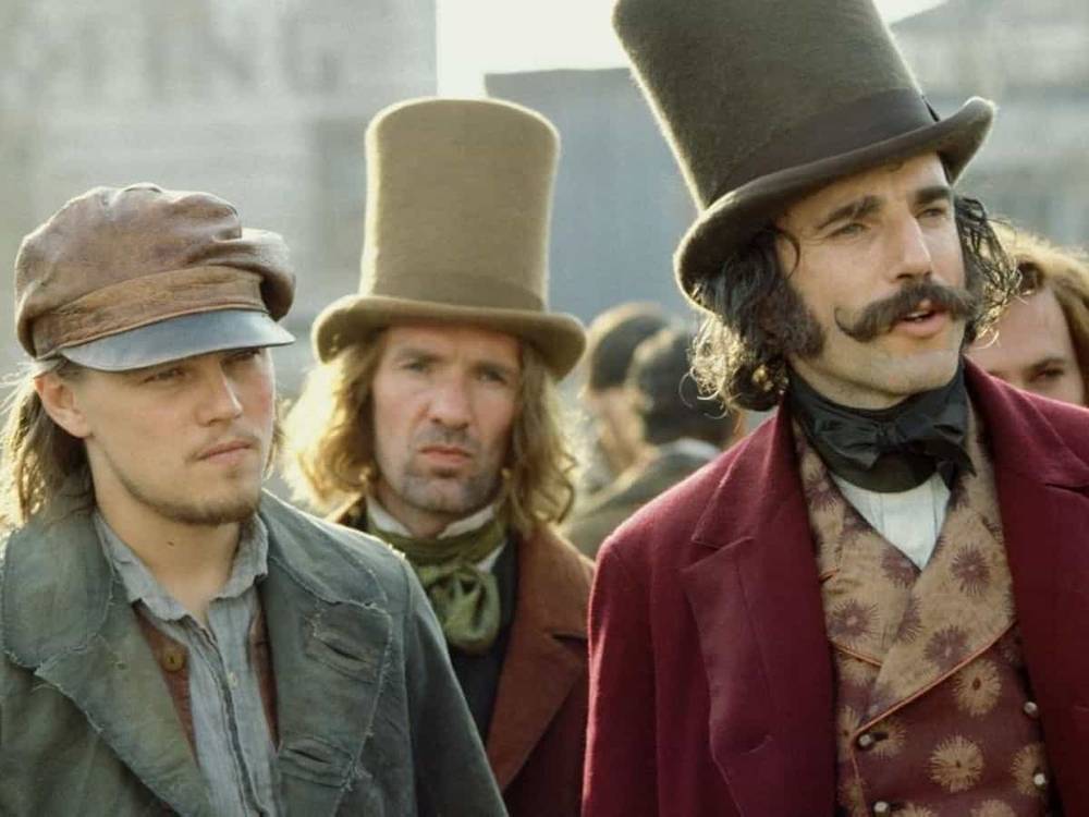 Trois hommes en tenue historique, deux portant des chapeaux hauts et un avec une casquette, se tiennent ensemble, représentant les personnages du film d'époque "Gangs of New York". L'homme au centre arbore un