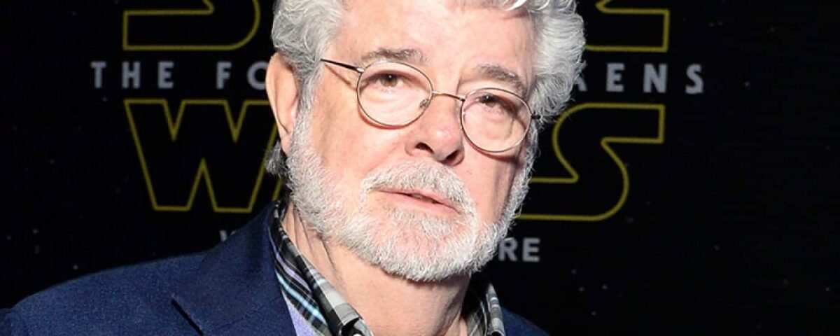 Un homme mûr aux cheveux et à la barbe blancs, ressemblant à George Lucas, portant une veste de costume sombre sur une chemise à carreaux, se tient devant un décor sur le thème de « Star Wars ».
