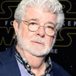 Un homme mûr aux cheveux et à la barbe blancs, ressemblant à George Lucas, portant une veste de costume sombre sur une chemise à carreaux, se tient devant un décor sur le thème de « Star Wars ».
