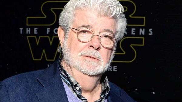 Un homme mûr aux cheveux et à la barbe blancs, ressemblant à George Lucas, portant une veste de costume sombre sur une chemise à carreaux, se tient devant un décor sur le thème de « Star Wars ».
