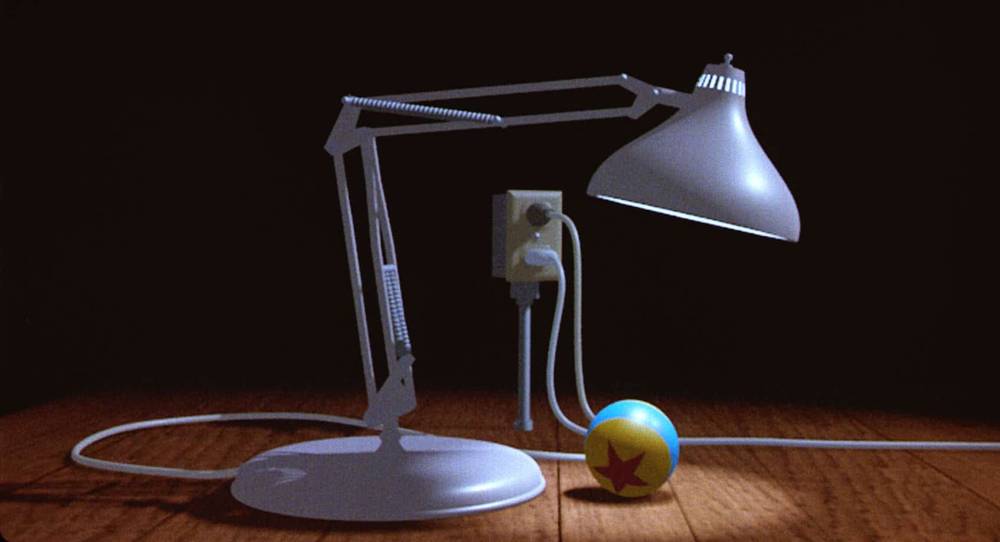 Une lampe Luxo animée se penche pour regarder une petite boule avec une étoile dessus, posée sur une surface texturée en bois dans une pièce faiblement éclairée.