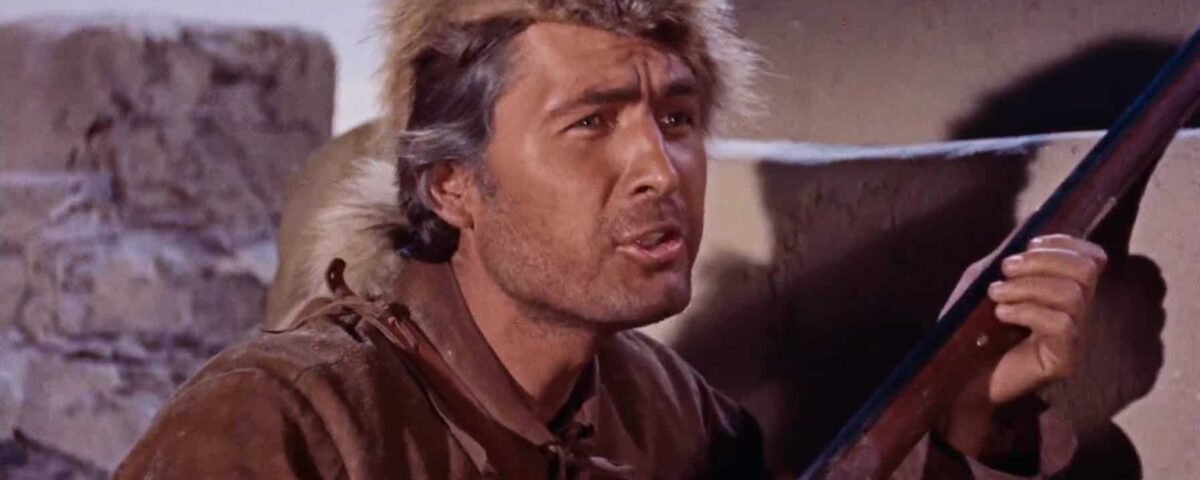 Un homme en costume historique de Davy Crockett, portant un chapeau de fourrure, lève les yeux avec inquiétude tout en tenant un fusil, sur fond de pierre.