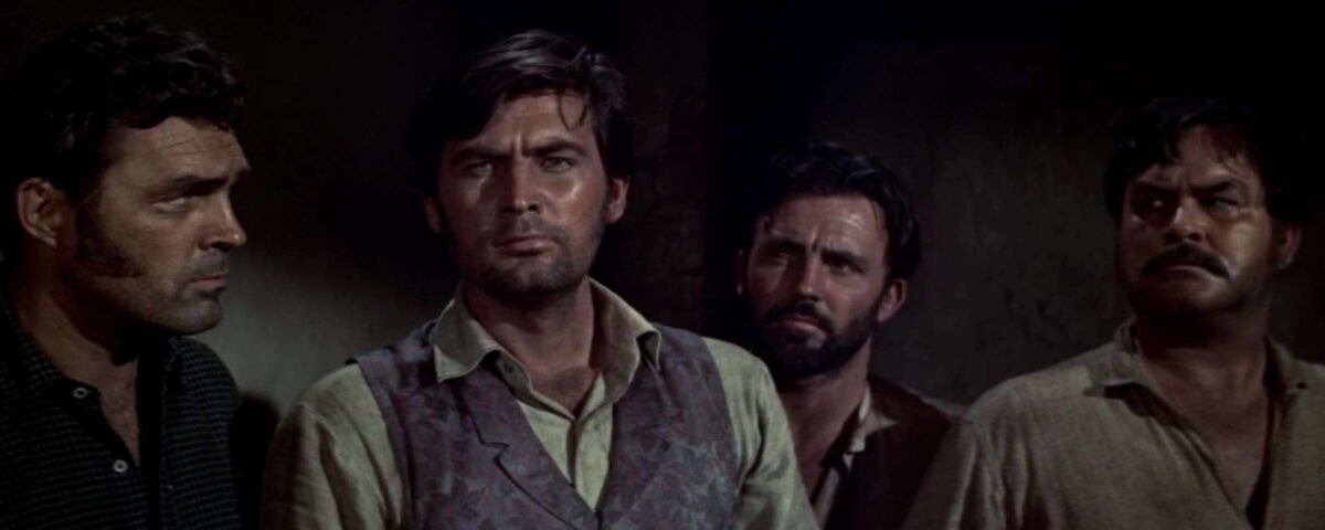 Quatre hommes aux expressions sérieuses se tiennent côte à côte, regardant de côté dans une pièce faiblement éclairée, tous vêtus de vêtements bruts et décontractés, comme s'ils anticipaient une poursuite infernale.