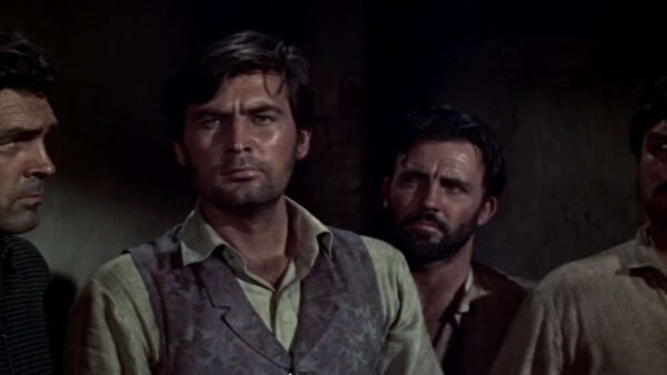 Quatre hommes aux expressions sérieuses se tiennent côte à côte, regardant de côté dans une pièce faiblement éclairée, tous vêtus de vêtements bruts et décontractés, comme s'ils anticipaient une poursuite infernale.