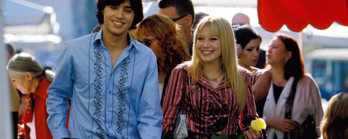 Un jeune homme et une jeune femme, rappelant une scène de Lizzie McGuire, sourient en se promenant dans un marché animé, l'homme tenant un tournesol. Ils sont habillés de façon décontractée et colorée