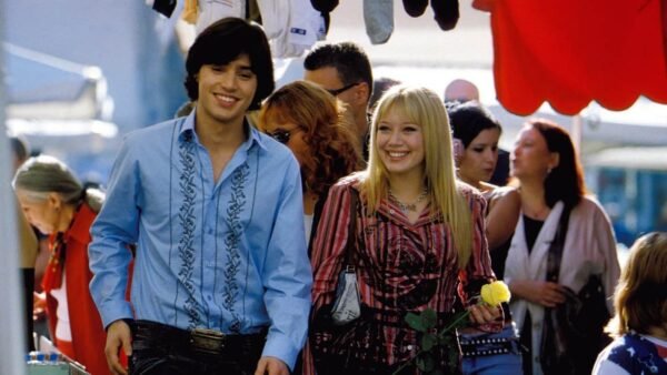 Un jeune homme et une jeune femme, rappelant une scène de Lizzie McGuire, sourient en se promenant dans un marché animé, l'homme tenant un tournesol. Ils sont habillés de façon décontractée et colorée