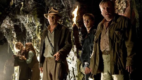 Trois hommes en tenue d'aventure se tiennent dans une grotte faiblement éclairée, tenant des torches, entourés de murs de pierre recouverts d'anciens glyphes, affichant des expressions de curiosité et d'appréhension alors qu'ils se préparent à entrer dans demain.