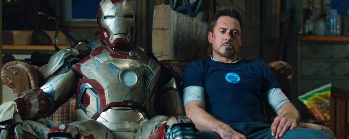 Tony Stark est assis avec lassitude à côté de son costume d'Iron Man dans un atelier encombré, reflétant un moment d'épuisement ou de contemplation.