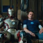 Tony Stark est assis avec lassitude à côté de son costume d'Iron Man dans un atelier encombré, reflétant un moment d'épuisement ou de contemplation.