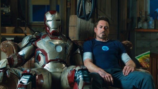 Tony Stark est assis avec lassitude à côté de son costume d'Iron Man dans un atelier encombré, reflétant un moment d'épuisement ou de contemplation.