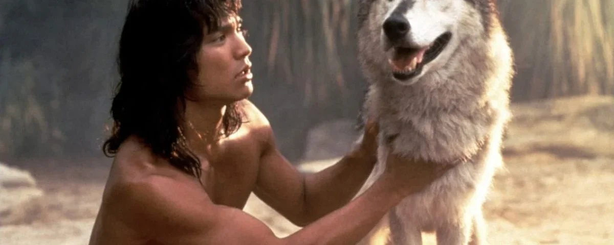 Un homme aux longs cheveux noirs, rappelant "Le Livre de la Jungle", vêtu d'une tenue minimale, posant dans un environnement sablonneux tout en tenant affectueusement par les épaules un grand loup souriant.