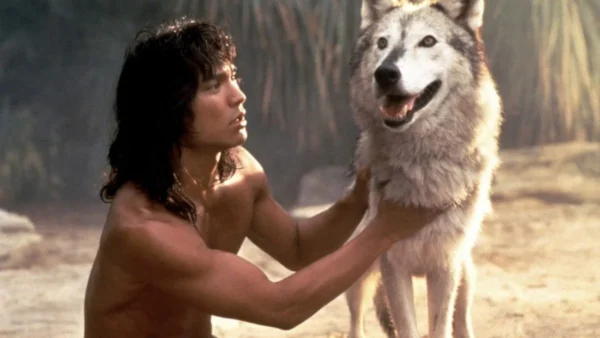 Un homme aux longs cheveux noirs, rappelant "Le Livre de la Jungle", vêtu d'une tenue minimale, posant dans un environnement sablonneux tout en tenant affectueusement par les épaules un grand loup souriant.