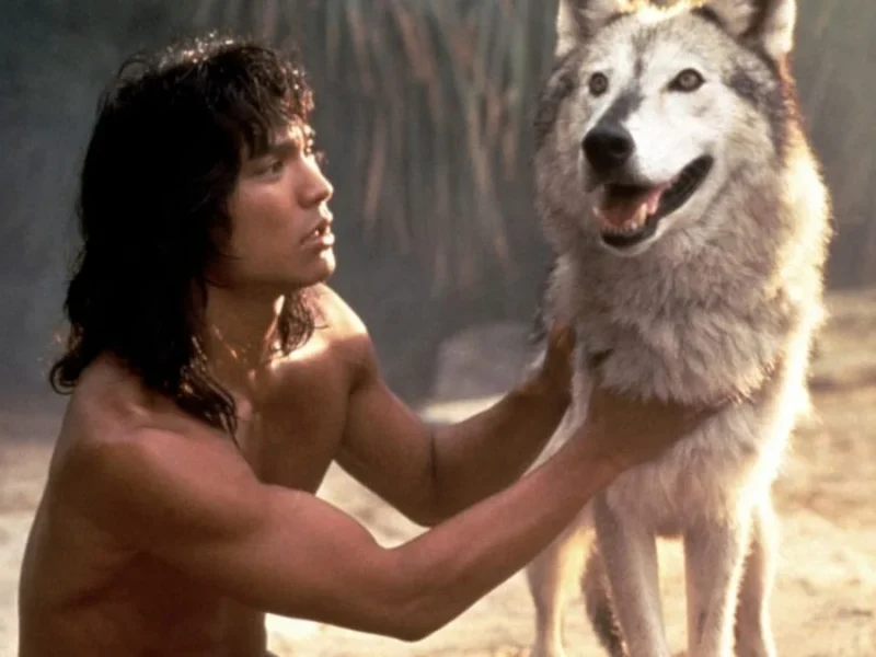 Un homme aux longs cheveux noirs, rappelant "Le Livre de la Jungle", vêtu d'une tenue minimale, posant dans un environnement sablonneux tout en tenant affectueusement par les épaules un grand loup souriant.