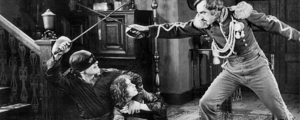 Un film en noir et blanc représentant une scène dramatique de combat à l'épée entre deux hommes en costumes ornés, avec une femme en détresse accroupie sur Main Street en arrière-plan.