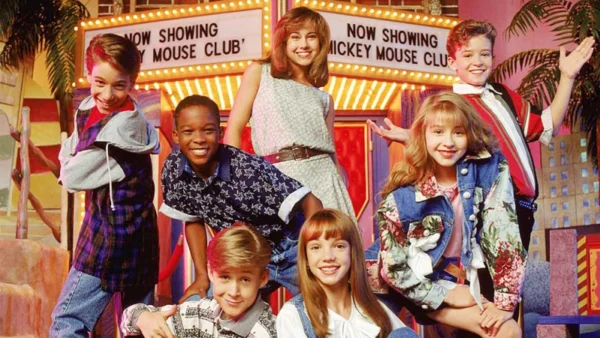Sept jeunes artistes sourient et posent sur le plateau du « Mickey Mouse Club », avec des vêtements colorés style années 90 et un fond sur lequel on peut lire « Now Showing Mickey Mouse Club ».