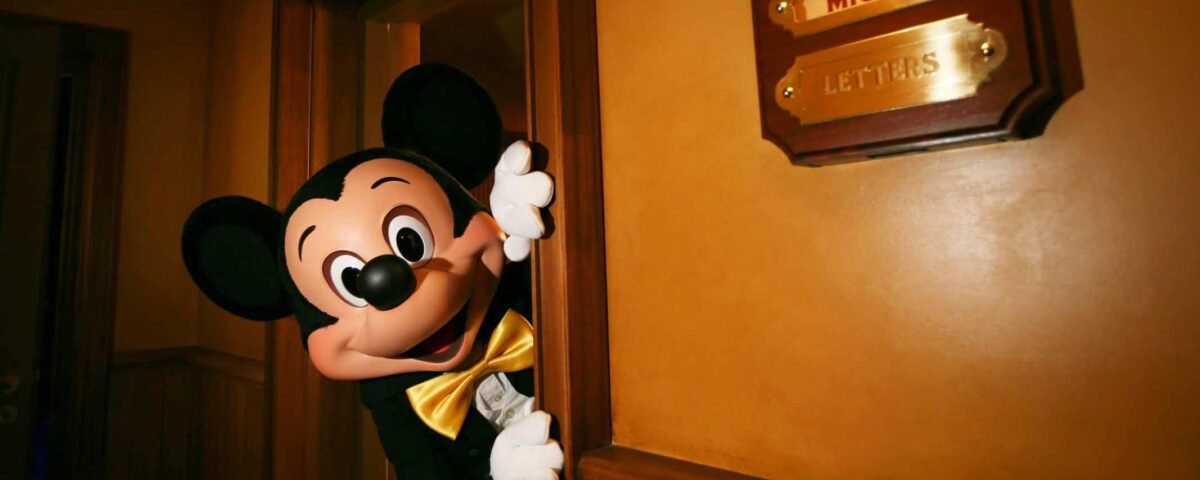 Mickey Mouse jette un coup d'œil derrière une porte légèrement ouverte dans une pièce chaleureusement éclairée de Fantasyland, à côté d'une boîte aux lettres murale intitulée "Lettres de Mickey".