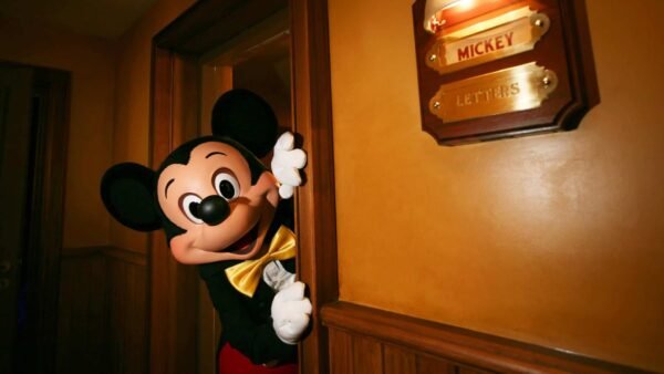 Mickey Mouse jette un coup d'œil derrière une porte légèrement ouverte dans une pièce chaleureusement éclairée de Fantasyland, à côté d'une boîte aux lettres murale intitulée "Lettres de Mickey".