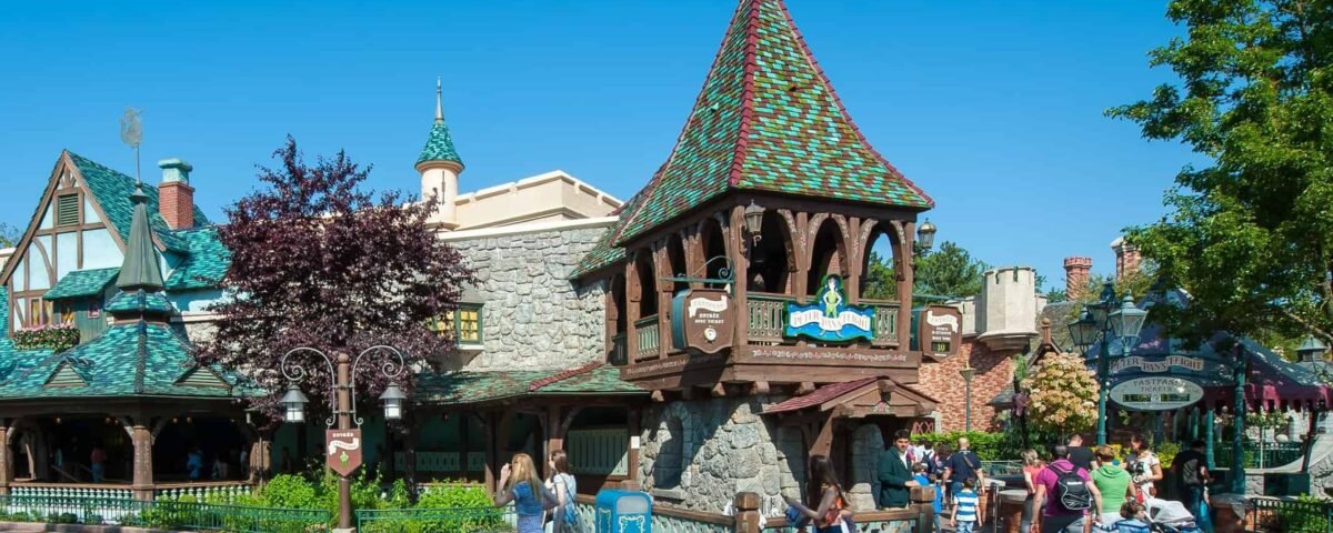 Les visiteurs se promènent dans un parc animé sur le thème de Peter Pan avec une architecture de conte de fées de style européen, avec un bâtiment en bois de premier plan avec un toit vert. Une journée lumineuse et ensoleillée renforce l’atmosphère joyeuse et animée.