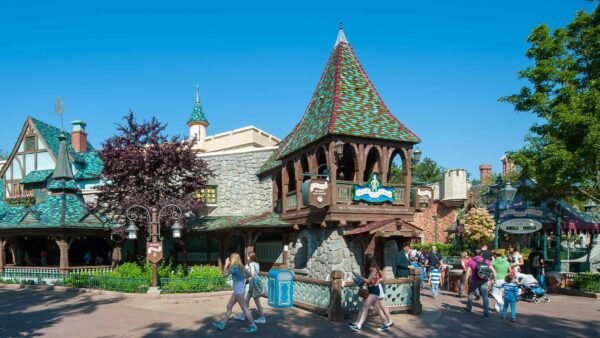 Les visiteurs se promènent dans un parc animé sur le thème de Peter Pan avec une architecture de conte de fées de style européen, avec un bâtiment en bois de premier plan avec un toit vert. Une journée lumineuse et ensoleillée renforce l’atmosphère joyeuse et animée.