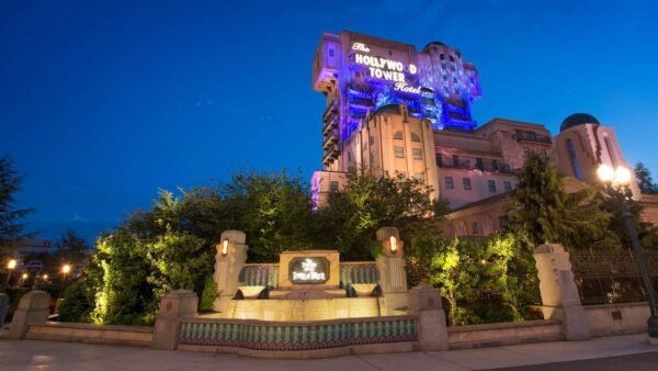 Vue au crépuscule de la Tour de la Terreur dans un parc à thème, avec une signalisation lumineuse sur la structure imposante et étrange, et un panneau d'entrée éclairé au premier plan.