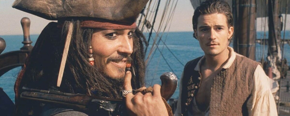 Un pirate des "Pirates des Caraïbes" avec un tricorne et des dreadlocks regarde pensivement sur le côté, tenant un petit télescope. A côté de lui se tient un jeune homme dans un