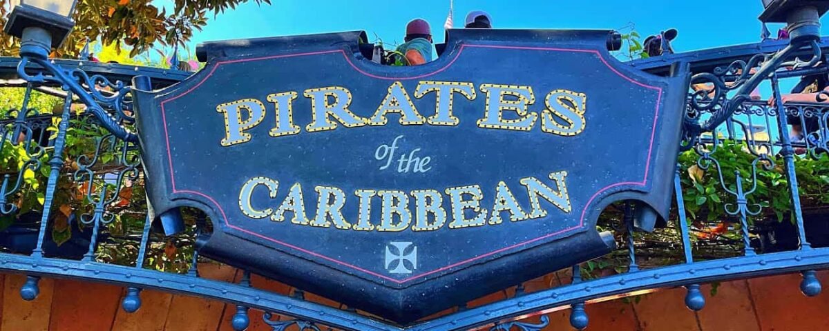 Panneau décoratif indiquant « Pirates des Caraïbes » avec des lettres dorées et ornées sur fond noir, sur un ciel bleu vif, encadré d'arbres verts feuillus.