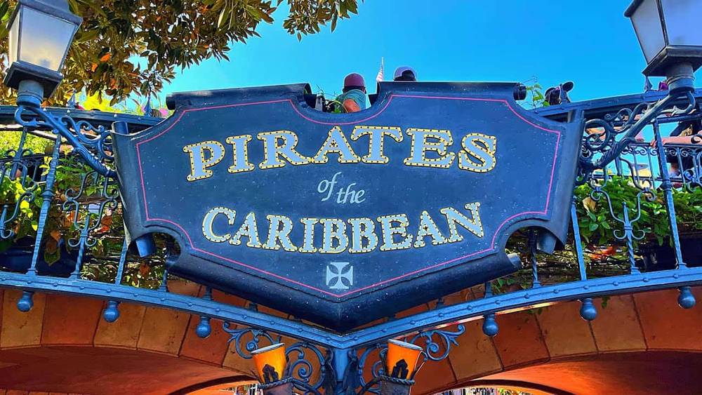 Panneau décoratif indiquant « Pirates des Caraïbes » avec des lettres dorées et ornées sur fond noir, sur un ciel bleu vif, encadré d'arbres verts feuillus.