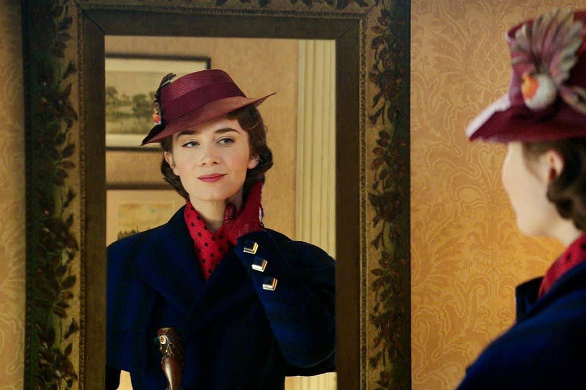 Une femme portant un chapeau marron vintage et un manteau bleu orné d'un foulard rouge regarde son reflet dans un miroir orné, ajustant son foulard avec une expression ravie rappelant "Mary Poppins Returns".