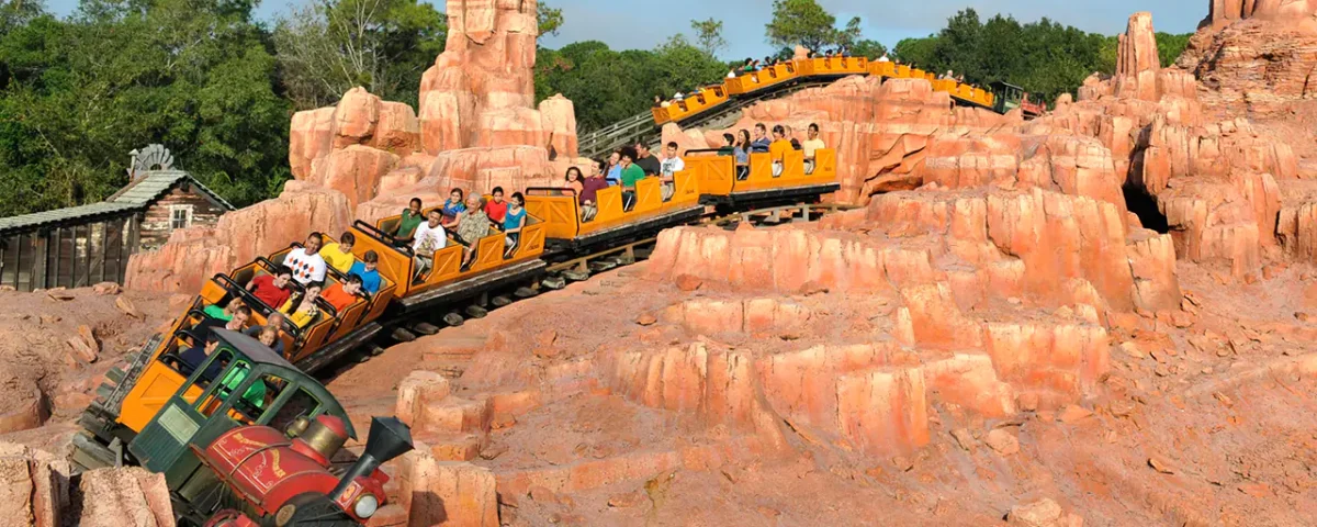 Des montagnes russes avec des passagers dans des chariots colorés roulant à toute vitesse le long d'une piste entourée d'un terrain rocheux artificiel sous un ciel clair, semblable à Big Thunder Mountain.