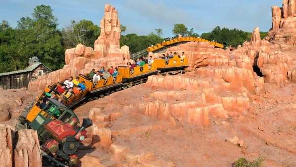 Des montagnes russes avec des passagers dans des chariots colorés roulant à toute vitesse le long d'une piste entourée d'un terrain rocheux artificiel sous un ciel clair, semblable à Big Thunder Mountain.