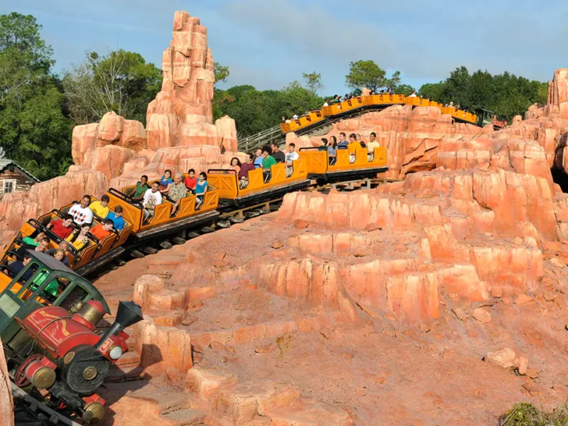 Des montagnes russes avec des passagers dans des chariots colorés roulant à toute vitesse le long d'une piste entourée d'un terrain rocheux artificiel sous un ciel clair, semblable à Big Thunder Mountain.