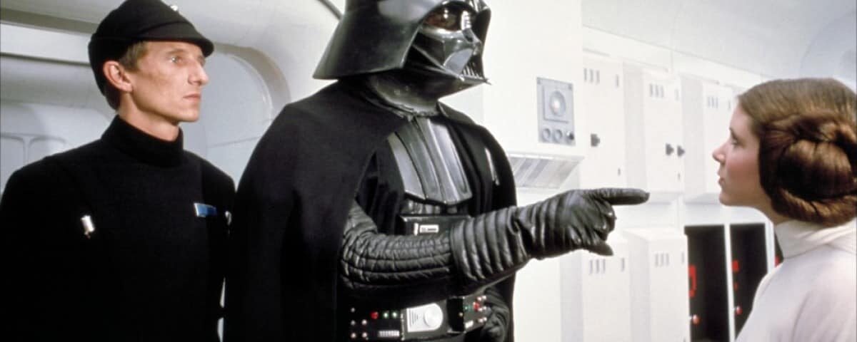 Dark Vador, portant son armure et son casque noirs, fait un geste de sa main gantée vers la princesse Leia, qui le regarde sévèrement, dans un couloir de vaisseau spatial d'un blanc éclatant dans "Un nouvel espo