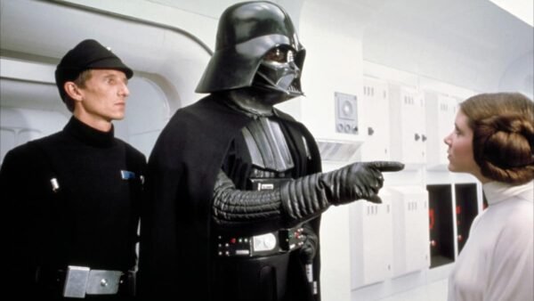 Dark Vador, portant son armure et son casque noirs, fait un geste de sa main gantée vers la princesse Leia, qui le regarde sévèrement, dans un couloir de vaisseau spatial d'un blanc éclatant dans "Un nouvel espo