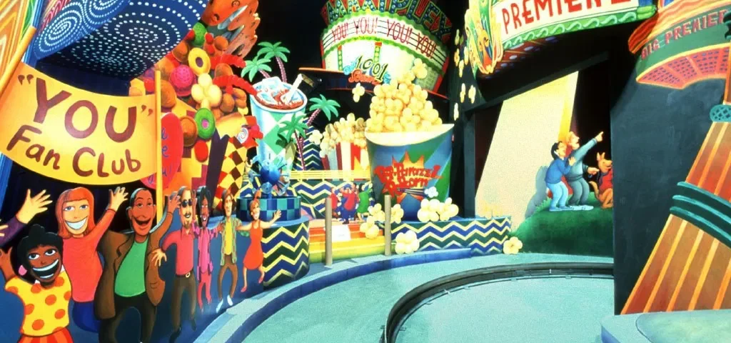 Parc à thème animé avec des personnages colorés et animés, dont une foule joyeuse et un clown, près du panneau « limousine superstar », sous une bannière « première ».