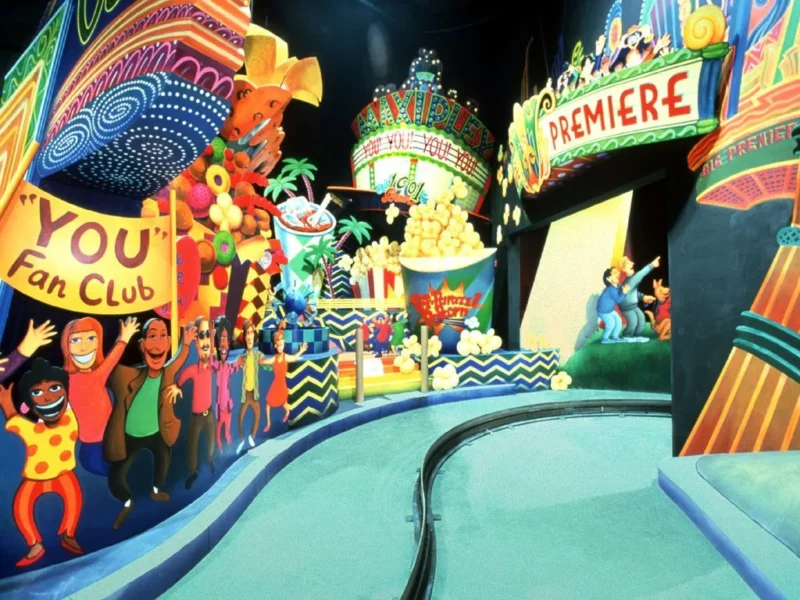 Parc à thème animé avec des personnages colorés et animés, dont une foule joyeuse et un clown, près du panneau « limousine superstar », sous une bannière « première ».