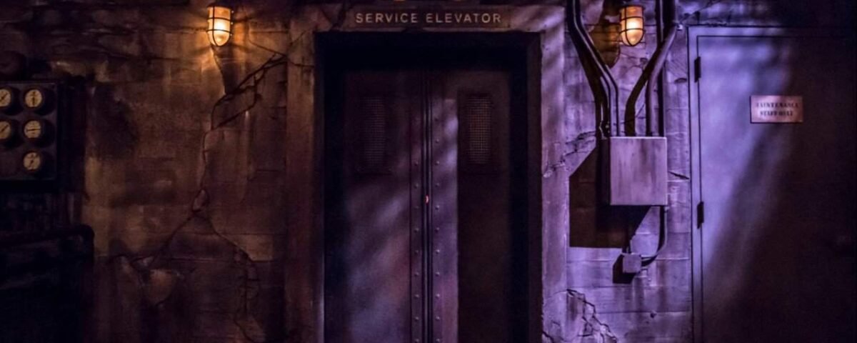 Une scène atmosphérique faiblement éclairée montrant une vieille porte d'ascenseur de service entourée de murs en pierre fissurés dans la Tour de la Terreur, avec un panneau indiquant "ascenseur de service" et un éclairage d'ambiance provenant de lampes murales.