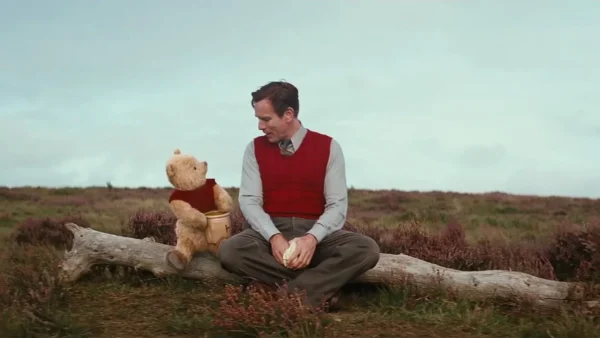 Un homme et un ours en peluche, Jean-Christophe Robin, sont assis sur une bûche en plein champ. L'homme, vêtu d'un pull et d'une cravate, apparaît contemplatif, tandis que l'ours a l'air contemplatif.