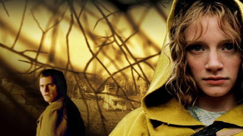 Affiche promotionnelle pour un film dramatique mettant en scène au premier plan une jeune femme en imperméable jaune, à l'air sombre, avec un homme flou et une scène de banlieue en arrière-plan du village.