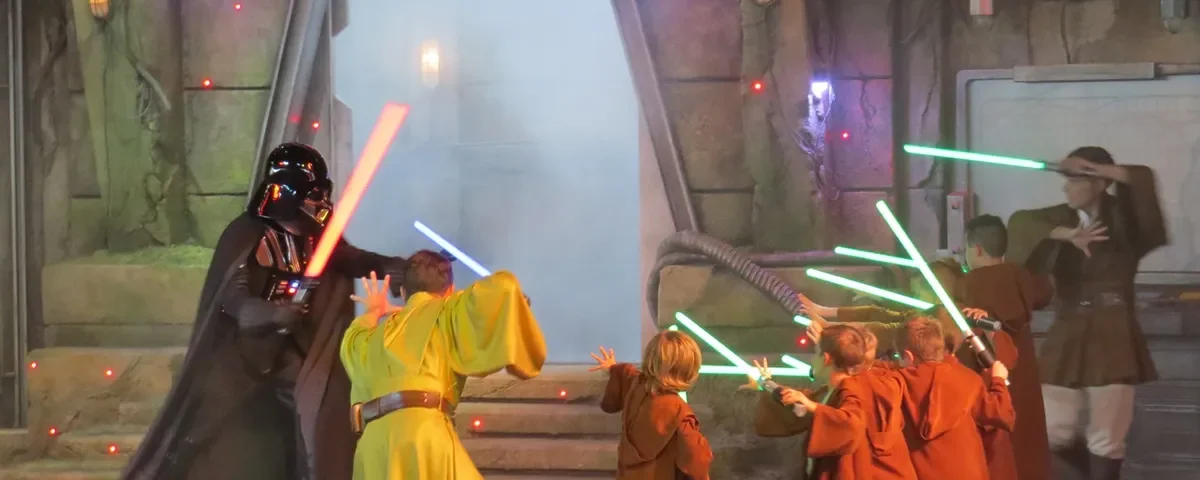 Des enfants habillés en stagiaires de l'Académie de formation Jedi tenant des sabres laser affrontent Dark Vador lors d'un événement sur le thème de Star Wars avec un fond enfumé et un éclairage théâtral.